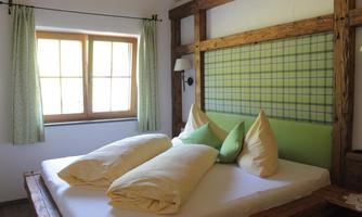 A cozy bedroom with a large bed and soft pillows. The walls are decorated with wood and a checkered pattern, and light streams through the windows.
