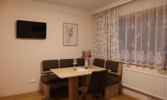 A cozy dining area with a wood-colored table and a bench-style seating. A TV and a framed picture are hanging on the wall.