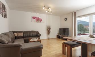 A modern living room with a large brown leather sofa and a dining table. The walls are bright, and there are windows overlooking the landscape.