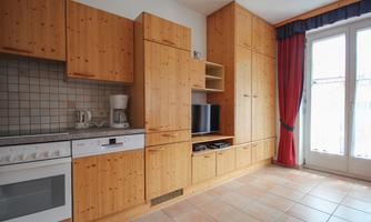 A modern kitchen with wooden cabinets and tiled floor. The room has a television and large windows with curtains.