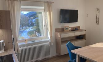 A cozy living space with a window overlooking a picturesque landscape. A television and a table with blue chairs complete the furnishings.