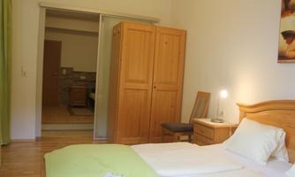 A bright bedroom with a double bed and a wooden wardrobe. In the background, another room is visible, which is comfortably furnished.