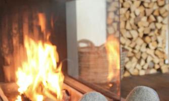 A cozy fireplace with a roaring fire and a stack of wood in the background. Visible are feet in warm slippers relaxing in front of the fireplace.