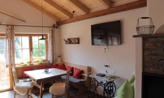 A cozy living space with a wooden ceiling and a large table. A television hangs on the wall, and there are colorful cushions on the seats.