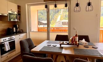 A modern kitchen dining area with a large table and comfortable chairs. Large windows let in plenty of light and offer a view of the mountains.