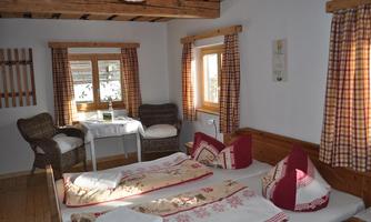 A cozy bedroom with a wooden ceiling and two beds. There is a small table with chairs and large windows with checkered curtains.