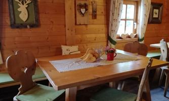 A cozy wooden room with a table and chairs that have green cushions. Decorative pictures hang on the wall, and a window lets in natural light.