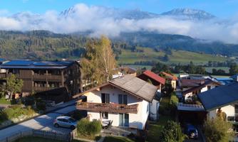 A picturesque village with modern buildings set against an impressive mountain landscape. The sky is partly cloudy, and the landscape is green and inviting.