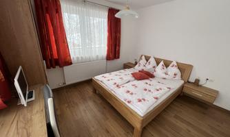A cozy bedroom with a double bed and red curtains. Next to the bed is a wooden desk with a chair.