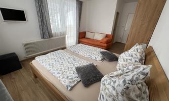A cozy bedroom with a double bed and an orange sofa. Large windows let in plenty of daylight.