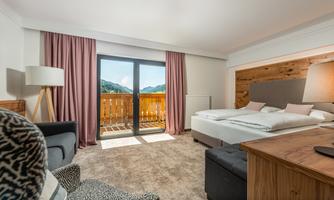A modern hotel room with a large window and a view of the mountains. It is cozily furnished with a bed, an armchair, and a table.