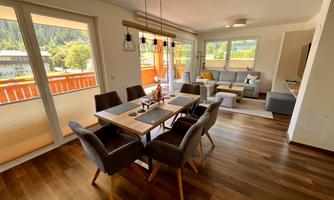 A modern living and dining room with a large dining table and cozy sofa. Large windows offer a view of the surrounding landscape.