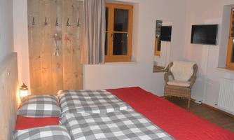 A cozy bedroom with a large bed and checkered bedspreads. Some coat hooks hang on the wall, and there is a television as well as a chair in the corner.