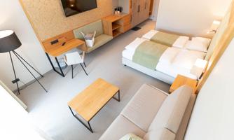 A modern hotel room with two beds, a sofa, and a table. The room is bright and friendly with a television on the wall.