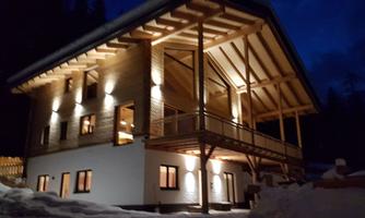 A modern wooden house at night, illuminated by warm lights. It is surrounded by snow and encompassed by a dark sky.