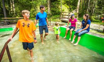 A family is playing in the shallow water in a natural setting. The children are having fun while the adults sit on a bench and watch.