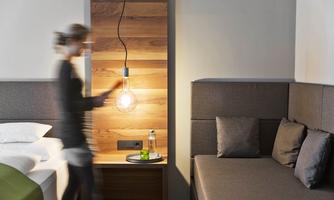 A modern hotel room with a bed and a cozy seating area. A person moves quickly in the room while a stylish lamp is shining.