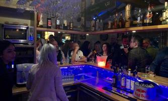 A lively bar with several guests and a variety of drinks behind the counter. The atmosphere is vibrant and inviting, with colorful lights and modern decor.