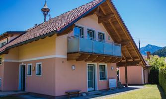 A beautiful house with a balcony and a red tiled roof. In the background, mountains and a blue sky can be seen.
