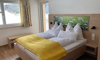 A cozy bedroom with a double bed and a yellow blanket. Large windows let in plenty of daylight and offer a view outside.