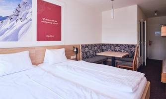 A bright room with two single beds and a table. Pictures of mountains hang on the wall, and a red wall informs about the hotel offers.