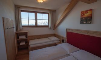 A cozy bedroom with two single beds and a large window. Snow-capped mountains are visible in the background.