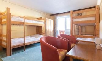 A simple room with two bunk beds and a table. Two red chairs are next to the table, and there is a window with a view outside.