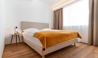 A bright bedroom with a double bed and an orange blanket. The room has large windows with curtains and a small table with a lamp.