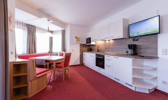 A modern kitchen corner with a dining table and red chairs. The kitchen is bright and well-equipped, with a TV on the wall.