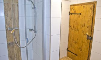 A modern bathroom with a shower and a classic wooden door. The wall is covered with white tiles and the floor is made of wood.
