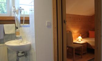A simple bathroom with a sink and a window. In the background, a cozy bedroom with a nightstand and a lamp is visible.