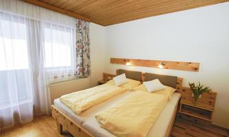 A cozy bedroom with a double bed and light wood decor. The curtains are airy and provide a view outside.