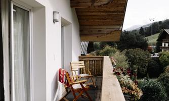 A quiet balcony with wooden flooring and two chairs. In the background, green areas and mountains are visible.