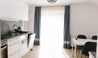 A bright and modern kitchen unit with white cabinets and a dining table. Large windows let in plenty of light and give the room an inviting atmosphere.