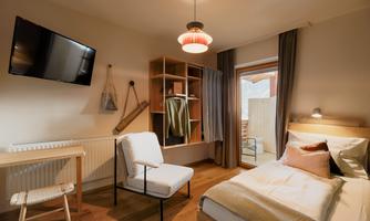 A modern room with a comfortable chair and a television. Large windows provide access to a balcony and ample daylight.