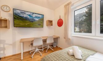 A modern room with a cozy bed and a desk. A large TV hangs on the wall and a window offers a view of the mountains.