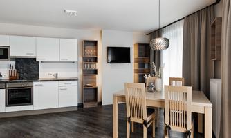A modern kitchen with a wooden dining table and four chairs. The atmosphere is bright and friendly, with large windows and minimalist decor.