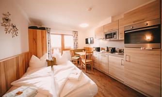 A cozy room with a double bed and wooden-paneled walls. There is a small kitchen and a dining area with a table and chairs.