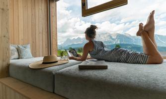 A woman is lying relaxed on a bed and reading a book. The view from the window reveals an impressive mountain landscape under a cloudy sky.