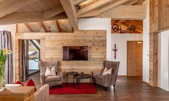 A cozy living room with wood paneling, two beige armchairs, and a small table. A television hangs on the wall, and there is a red carpet on the floor.