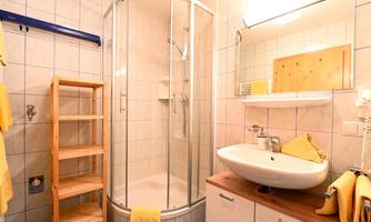 A modern bathroom with a shower and a sink. The walls are tiled and there are wooden shelves as well as yellow towels.