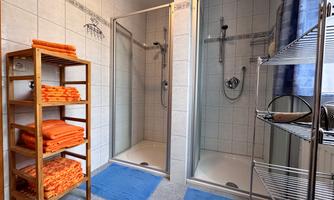 A modern bathroom with two shower cabins and a shelf with orange towels. The floor is covered with blue mats.