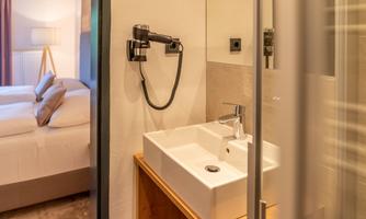 A modern bathroom with an elegant sink unit. In the background, a bright bedroom and a shower can be seen.