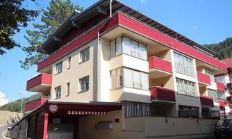 A modern multi-family house with red and white facade design. It is located in a quiet environment, surrounded by trees and mountains.