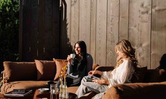 Two women are sitting relaxed on the designer couch in the garden of the ARX Hotel, enjoying their time together. One of them is holding a cup while the other is looking at a book.
