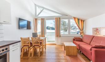 A bright and modern living and dining room with a red sofa and a wooden table. Large windows provide plenty of natural light and a pleasant view outside.