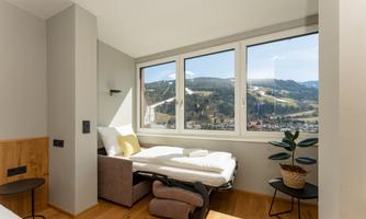 A bright bedroom with a large window and a view of the mountains. The room features a bed, a table, and a plant.