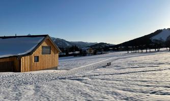 A picturesque winter landscape with a wooden house and a snow-covered meadow. The mountains are visible in the background and the sky is clear and blue.