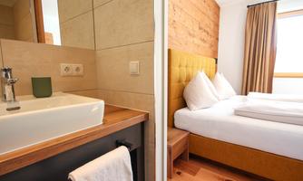 A modern room with a comfortable bed and a bright, friendly atmosphere. In the background, a stylish bathroom with a beautiful sink can be seen.
