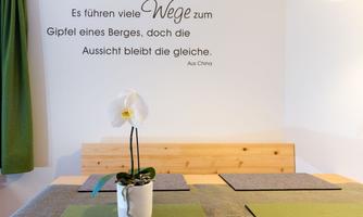 A cozy room with a wooden table and a white orchid in a pot. A quote is placed on the wall that deals with the path to the summit.